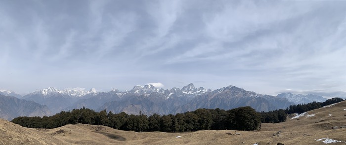 wide shot of Himalayan range. a valley with a tree line with the mountains in the back and the wide blue sky overheard